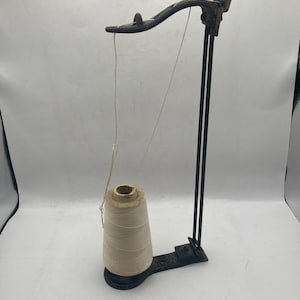 Puede incluir: Un portahilos metálico antiguo con un carrete de hilo beige. El portahilos es de color marrón oscuro y tiene un brazo curvado y soportes verticales. El carrete tiene forma de cono y se asienta sobre una base negra.