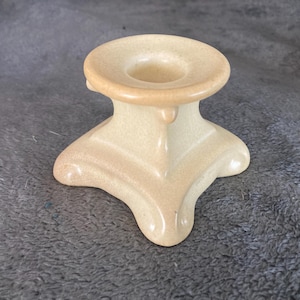 May include: A ceramic candle holder with a square base and a round top. The holder is a light beige color.