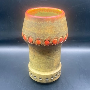 May include: A vintage ceramic vase with a unique shape and design. The vase features a wide top with a reddish-orange rim, transitioning to a yellow-brown body. It has a series of orange circles around the top and a row of holes near the base. The vase has a textured surface.