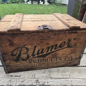 May include: A rectangular wooden crate with a hinged lid and a metal latch. The front panel features black printed text that reads Blumer PRODUCTS CO MONROE WIS. The wood has a weathered brown finish with visible grain and metal corner reinforcements.