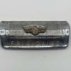 Pode incluir: Emblema Frigidaire vintage fabricado pela General Motors. O emblema retangular é prateado com uma textura canelada e um logotipo central. A palavra "FRIGIDAIRE" é exibida em letras maiúsculas.