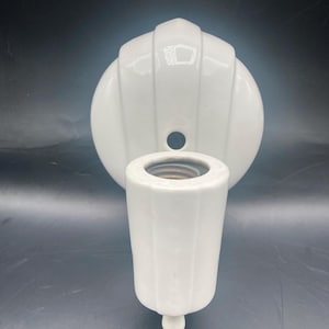 May include: A white ceramic light fixture with a cylindrical socket and a decorative backplate. The backplate has a rounded shape with vertical ridges. The socket has a small, rounded base. The fixture is designed for a light bulb.