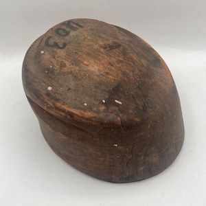 May include: A vintage, oval-shaped wooden hat block with visible wood grain and cracks. The block is a dark brown color with the number "403" stenciled in black on the top. Small white spots are scattered across the surface.