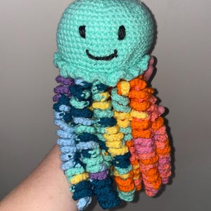 May include: Handmade crochet jellyfish toy in shades of teal, blue, purple, yellow, orange, and pink. The jellyfish has a round head with a smiling face and colorful tentacles. A whimsical and unique handmade toy.