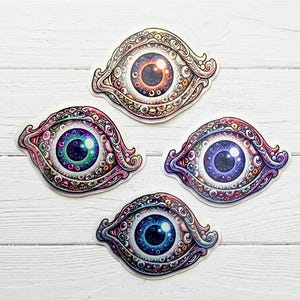 May include: Four die-cut stickers featuring stylized eye designs. Each eye is surrounded by a complex pattern of smaller, interconnected eyes. The central irises are colored in shades of orange, green, purple, and blue, with white highlights.