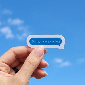 May include: A white sticker with a blue border and the text "Sorry, I was pooping" in a speech bubble shape. The sticker is held against a bright blue sky with scattered clouds.