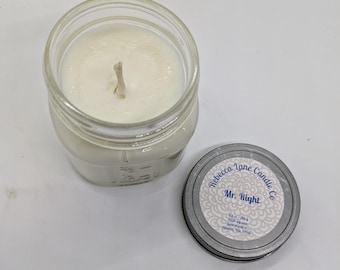 May include: A clear glass jar candle with a white wax interior and a wooden wick. The lid is off, revealing the candle's surface. The lid has a label that reads "Rebecca Lane Candle Co. Mr. Right."