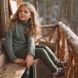 May include: A young person wearing a green turtleneck sweater and matching leggings sits on a wooden bench. The sweater has a white chevron pattern. The person wears burgundy shoes with white soles. The background is a forest.