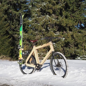 May include: A wooden bicycle with a brown leather seat and black tires is parked in the snow. A pair of green and black skis are attached to the bike's rack.