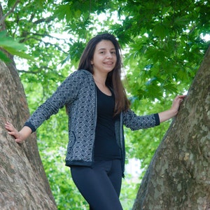 May include: A woman wearing a black top, black leggings, and a gray cardigan with a white wave pattern. She is standing between two tree trunks, with green leaves in the background.