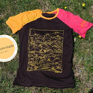 以下が含まれることがあります： 黒いラグランTシャツ、袖は黄色とピンク。山脈の黄色のグラフィックと「Himalaya Creative」の文字が特徴です。左側の円形グラフィックには「custom-made for you」と書かれています。