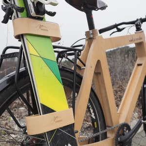 May include: A wooden bicycle with a pair of skis attached to the front. The skis are secured with a wooden strap that has the word "MONACO" printed on it. The bicycle is made of natural wood and has a black seat and handlebars.