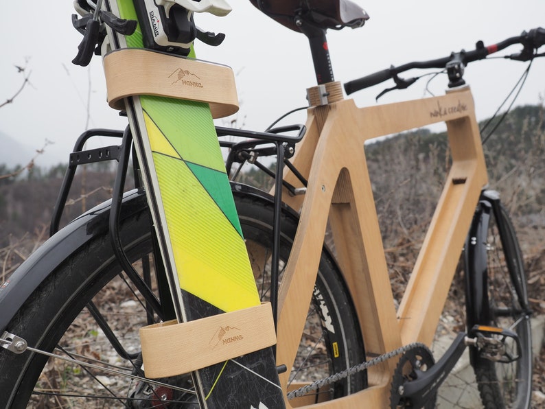 May include: A wooden bicycle with a pair of skis attached to the front. The skis are secured with a wooden strap that has the word "MONACO" printed on it. The bicycle is made of natural wood and has a black seat and handlebars.
