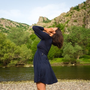 May include: A navy blue dress with long sleeves and a flared skirt. The dress has a gold zipper detail on the side. The background features a river, trees, and a mountain range.