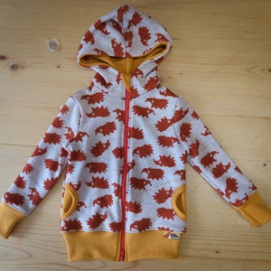 May include: A children's zip-up hoodie with a light gray background and a repeating pattern of orange hedgehog illustrations. The hoodie features a bright orange interior, cuffs, and pocket trim, with a red zipper.