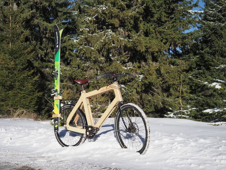 May include: A wooden bicycle with a brown leather seat and black tires is parked in the snow. A pair of green and black skis are attached to the bike's rack.