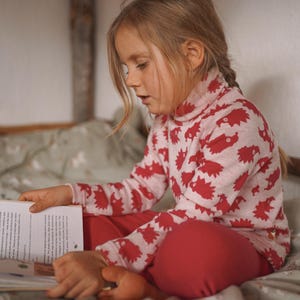 May include: A young girl wearing a pink and red long-sleeved top with a hedgehog print sits on a bed and reads a book.