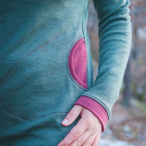 Op de afbeelding: Een teal-groene jurk met lange mouwen en een grote, afgeronde zak in een contrasterende magenta kleur. De manchetten van de mouwen zijn ook magenta. De jurk heeft decoratieve stiksels in magenta, wat een uniek designelement toevoegt.