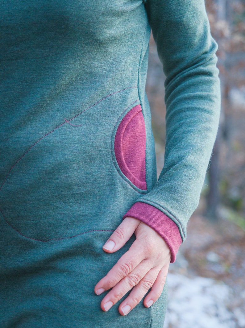 Op de afbeelding: Een teal-groene jurk met lange mouwen en een grote, afgeronde zak in een contrasterende magenta kleur. De manchetten van de mouwen zijn ook magenta. De jurk heeft decoratieve stiksels in magenta, wat een uniek designelement toevoegt.