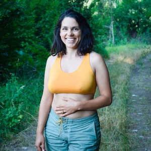 Puede incluir: Una persona con un sujetador deportivo naranja y pantalones cortos azul claro está de pie en un camino forestal. El sujetador deportivo es de color naranja brillante. Los pantalones cortos son azul claro con cordón.