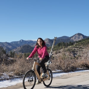 May include: A woman in a pink jacket rides a wooden bicycle with skis strapped to the back. She is smiling and looking at the camera. The background is a mountain range with snow-capped peaks.