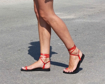 Può includere: Un paio di sandali in pelle rossa con sottili cinturini che si legano alle gambe. I sandali hanno una suola piatta e sono indossati da una donna.