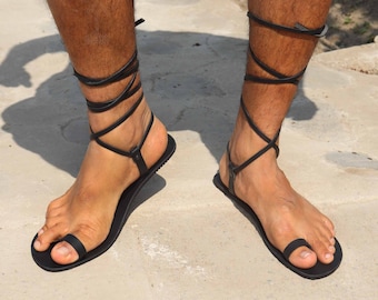 May include: Black leather sandals with laces that wrap around the ankles. The sandals have a flat sole and a toe loop.