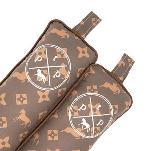 May include: Two brown rectangular door stops with a repeating horse and floral pattern. Each door stop features a white circular emblem with a horse silhouette and the letters "p" and "t". The door stops have brown trim and a fabric loop.