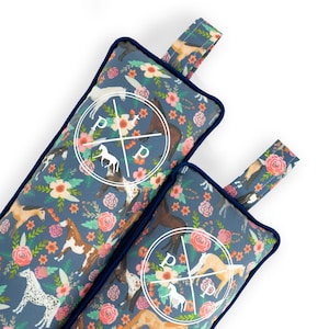 May include: Two rectangular fabric pouches with a dark blue background featuring a repeating pattern of various horses, pink flowers, and green leaves. Each pouch has a navy blue piped edge, a fabric loop at the top, and a white circular logo.