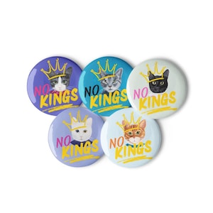 Puede incluir: Cinco botones redondos con ilustraciones de gatos con coronas. Cada botón tiene las palabras "NO KINGS" en amarillo. Los gatos son de varios colores, incluyendo negro, gris y naranja, sobre fondos morados, azules y blancos.