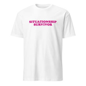 May include: A white t-shirt featuring the words "SITUATIONSHIP SURVIVOR" in vibrant pink lettering across the chest. The tee has a crew neck and short sleeves.