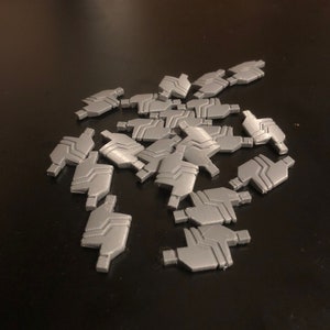 Puede incluir: Un montón de piezas impresas en 3D de plástico gris con un diseño geométrico. Las piezas tienen forma de un pequeño bloque rectangular plano con un borde curvo.