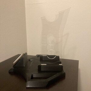 Pu&ograve; includere: Un supporto in stampa 3D nero con un supporto per telefono in plastica trasparente. Il supporto ha una fessura per un telefono e un'area rialzata per la ricarica.