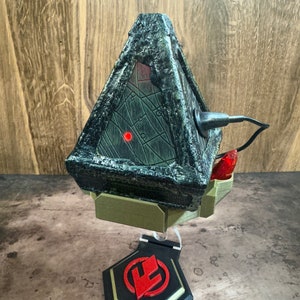 Può includere: Un oggetto di colore verde scuro, di forma triangolare, con una luce rossa e un filo nero. L'oggetto è su un supporto dorato con una base nera e un logo rosso.
