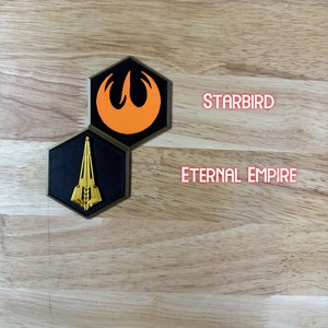 May include: Two hexagonal patches with embroidered logos. The top patch is orange and black with a stylised bird design. The bottom patch is black with a gold spaceship design. The text "STARBIRD" is above the top patch and "ETERNAL EMPIRE" is below the bottom patch.