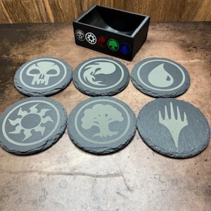 May include: Six black slate coasters with different Magic: The Gathering mana symbols in a light gray color. The symbols are: black, blue, red, green, white, and colorless. A black wooden coaster holder with the same symbols is in the background.