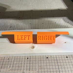 May include: Two orange plastic pieces with the words "LEFT" and "RIGHT" printed on them. The pieces are sitting on a wooden surface with a grid pattern.