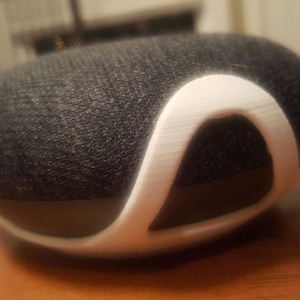 Op de afbeelding: Een zwarte en witte slimme speaker met een witte beschermende plastic hoes. De speaker heeft een stoffen bekleding en een witte plastic ring rond de bovenkant.