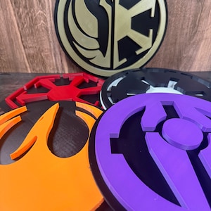 Könnte beinhalten: Fünf verschiedene Star Wars-Logos, jedes mit einer anderen Farbe und einem anderen Design. Die Logos sind aus Kunststoff und haben eine kreisförmige Form. Die Logos sind in einem Stapel angeordnet, wobei das oberste Logo ein goldenes und schwarzes Logo der Rebellenallianz und des Galaktischen Imperiums ist.