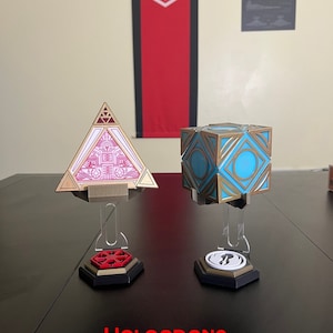 Può includere: Due supporti di visualizzazione stampati in 3D per gli ologrammi di Star Wars. I supporti sono in acrilico trasparente e presentano una base esagonale con un simbolo dell'universo di Star Wars. Il testo "HOLOCRONS NOT INCLUDED" è stampato in rosso nella parte inferiore dell'immagine.