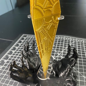 May include: A black 3D printed stand with four hands reaching up to hold a gold, geometric, 3D printed object. The text "STAND ONLY. LEVIATHAN NOT INCLUDED" is printed above the object.