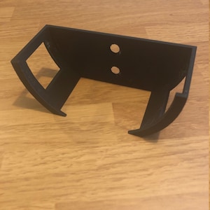 May include: Black plastic wall mount with two holes for mounting screws. The mount is designed to hold a cylindrical object, such as a speaker or a roll of paper towels.