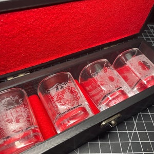 May include: A set of four clear glass shot glasses with intricate etched designs inside a black wooden box with a red velvet lining.