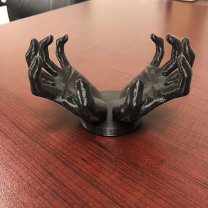 Peut inclure: Une sculpture noire imprimée en 3D représentant deux mains tendues vers le haut, montée sur une base circulaire. Les mains sont détaillées avec des doigts visibles et une surface texturée. La sculpture est placée sur un fond flou.