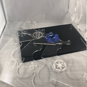 May include: A set of clear acrylic display stands featuring various Star Wars logos etched onto them. The stands are designed to hold miniature figures or other collectibles.