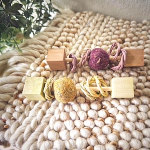 May include: Two horizontal pet chew toys featuring wooden cubes at each end, connected by a central rod. The top toy has a dark purple textured sphere and pink twisted fibers. The bottom toy has a yellow textured sphere and tan fibers.