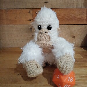 May include: A white crocheted gorilla plush toy sitting on a wooden surface with a large orange 20-sided die. The die has the numbers 14, 18, and 20 visible.