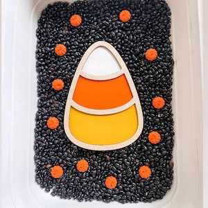 May include: A white plastic bin filled with black beans and small orange pumpkin-shaped figures surrounding a central wooden candy corn cutout. The candy corn features three horizontal sections colored white, orange, and yellow with a light wood frame.