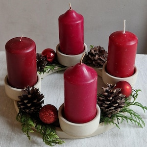 May include: A festive Advent wreath with four red candles, each in a cream-colored holder. The wreath is decorated with pinecones, red ornaments, and evergreen sprigs, creating a warm, seasonal display.