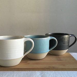 May include: Three ceramic mugs in different colors: white, light blue, and dark gray. The mugs have a rounded shape and a handle on the right side. They are arranged in a row on a wooden surface.